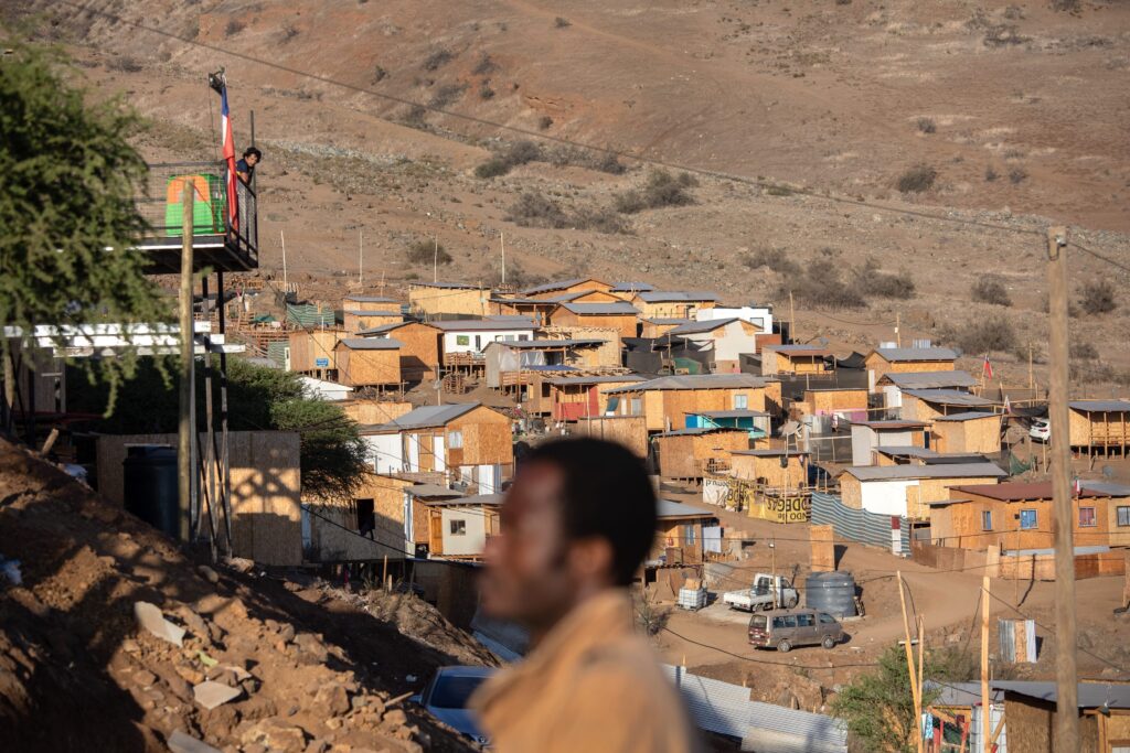 The out-of-focus profile of a man stands in the foreground while behind him in the distance lies a cluster of rudimentary shelters at the foot of a brown-colored hill dotted with tufts of brownish-green brush.