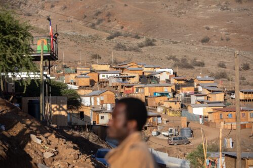 The out-of-focus profile of a man stands in the foreground while behind him in the distance lies a cluster of rudimentary shelters at the foot of a brown-colored hill dotted with tufts of brownish-green brush.