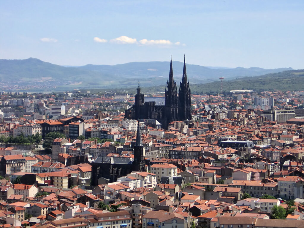 Two black church spires rise above buildings with red roofs, with mountains in the background under a blue sky.