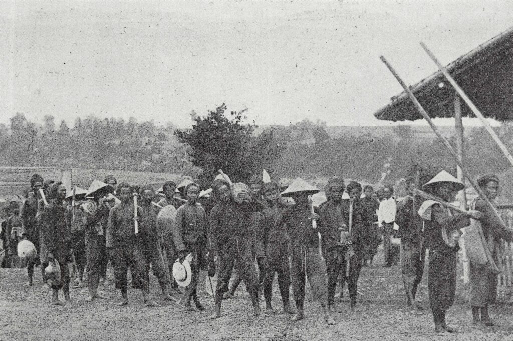 A grainy black-and-white photograph shows a group of manual laborers, some wearing peaked hats, standing outside next to a shelter made of wood poles and a roof.