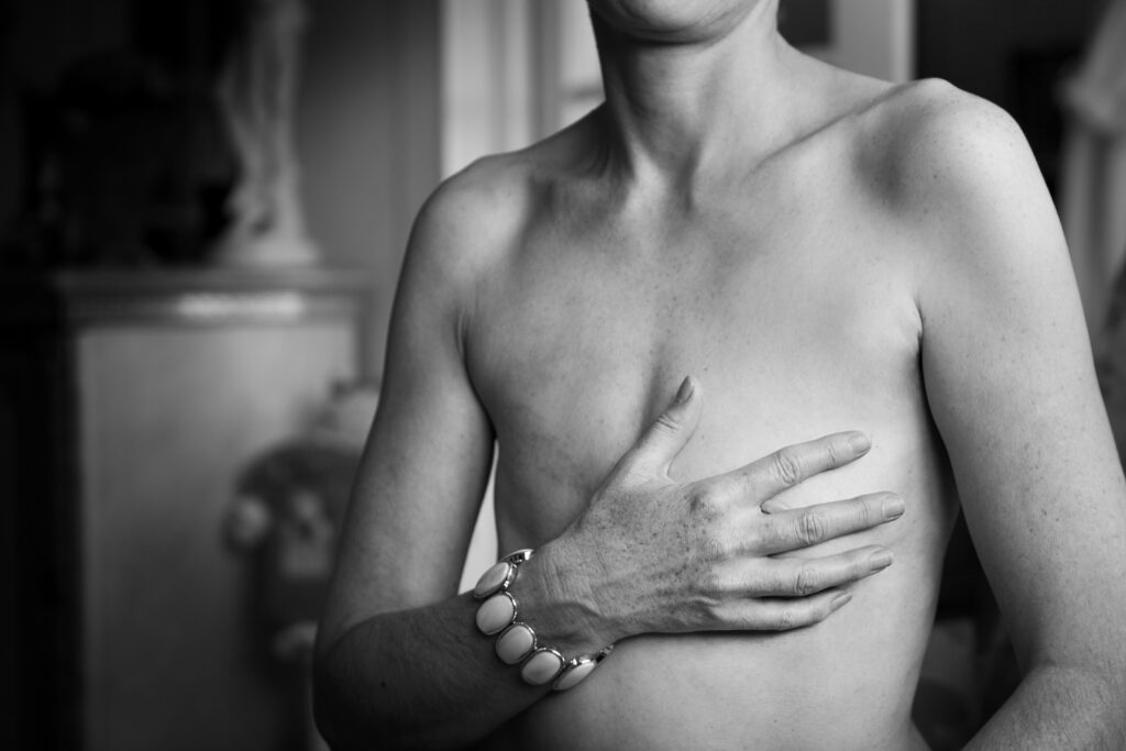 A black-and-white photograph shows a woman’s chest and torso. Her right hand, which wears a bracelet, reaches across her body to cover her chest.
