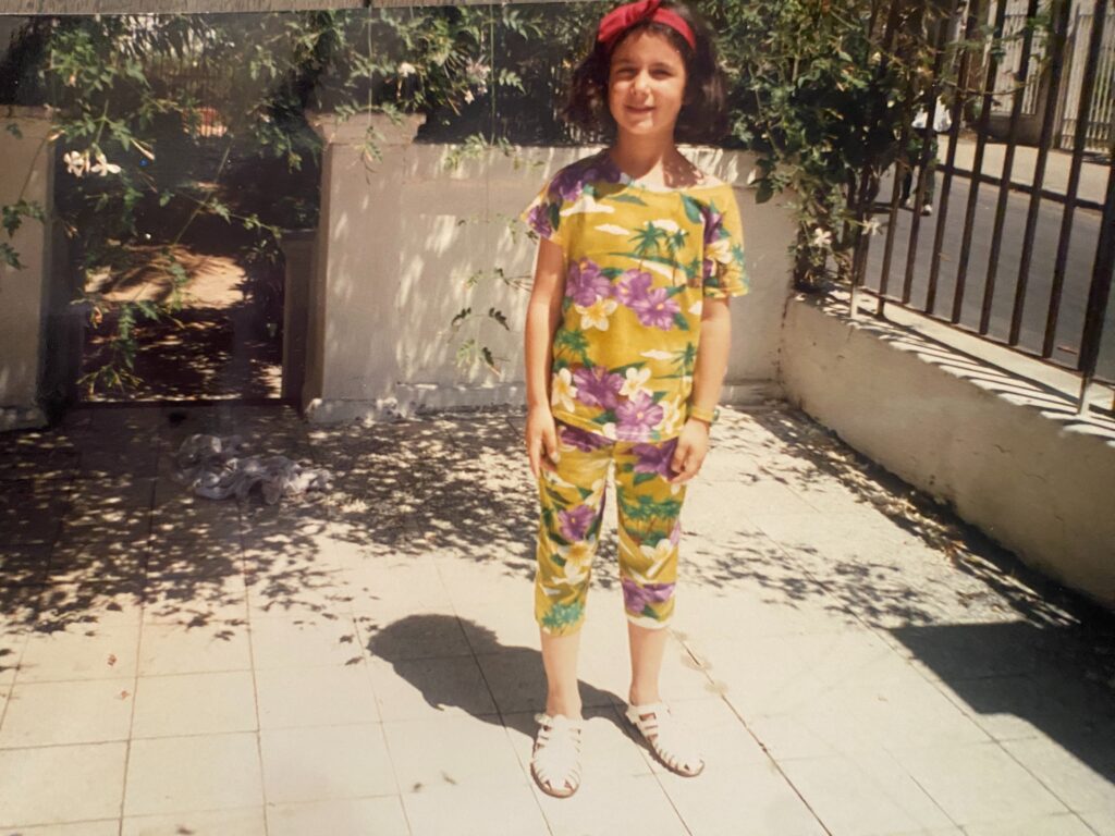 A young girl with a red bandana in her hair wearing a yellow outfit printed with tropical flowers and palm trees stands in the sunshine next to a wrought iron gate and tree branches with green leaves.