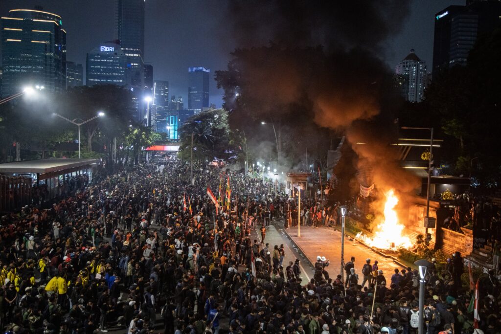 Amid tall city buildings lit up at night, a huge crowd gathers as smoke rises from a bonfire.
