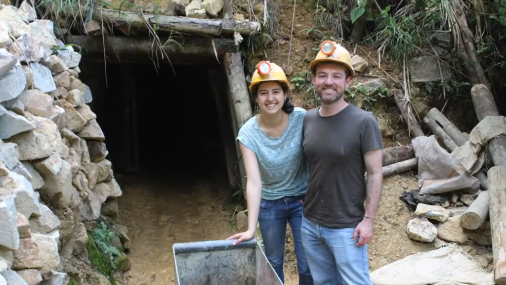 Duas pessoas usando capacetes amarelos com lanternas estão sorrindo, de braços dados, do lado de fora da entrada da mina, cercada por pedras e madeira.