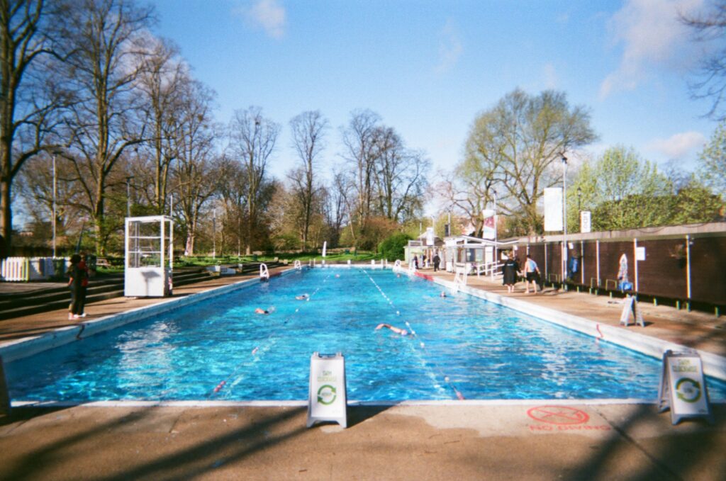Swimmers take laps in an outdoor swimming pool. On one side is a white sign on the cement deck and to the right are short structures for sitting and changing along the perimeter of the deck.