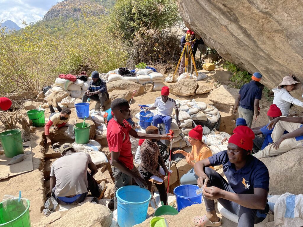Beside a large rock, a team digs at an archaeological site scattered with blue and green buckets and sandbags. Some team members wear red hats.