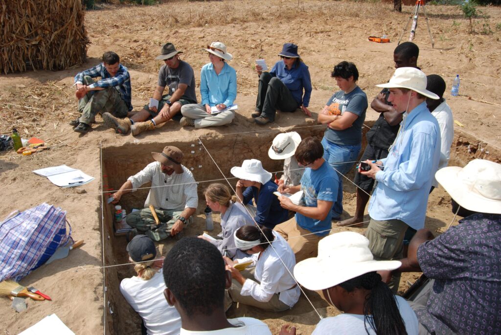 Sitting and standing within and around a square-shaped hole gridded with thin white strings, people look on and take notes as a person in a brown, broad-brimmed hat scrapes the inside wall of the hole.