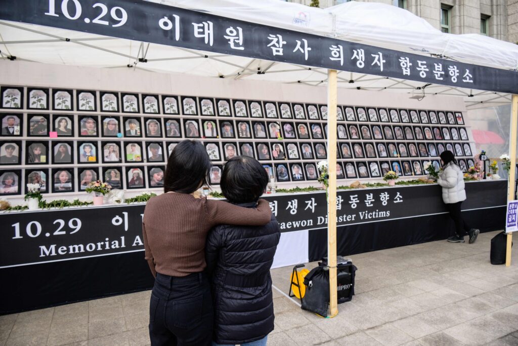 A woman with dark hair wearing a brown shirt wraps her arm around another woman with dark hair wearing a black coat as they look at a large memorial display of victims’ portraits. A black banner with white Korean letters appears above the display.