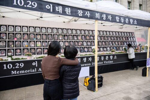 A woman with dark hair wearing a brown shirt wraps her arm around another woman with dark hair wearing a black coat as they look at a large memorial display of victims’ portraits. A black banner with white Korean letters appears above the display.