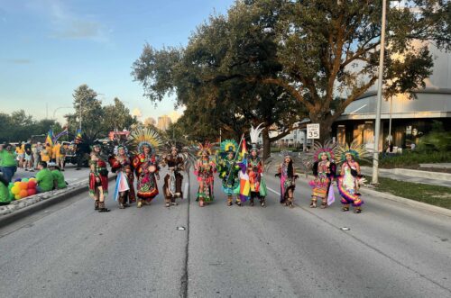 A group of people in elaborate bright-colored ceremonial clothing and headdresses stands in the middle of a tree-lined street in daylight.