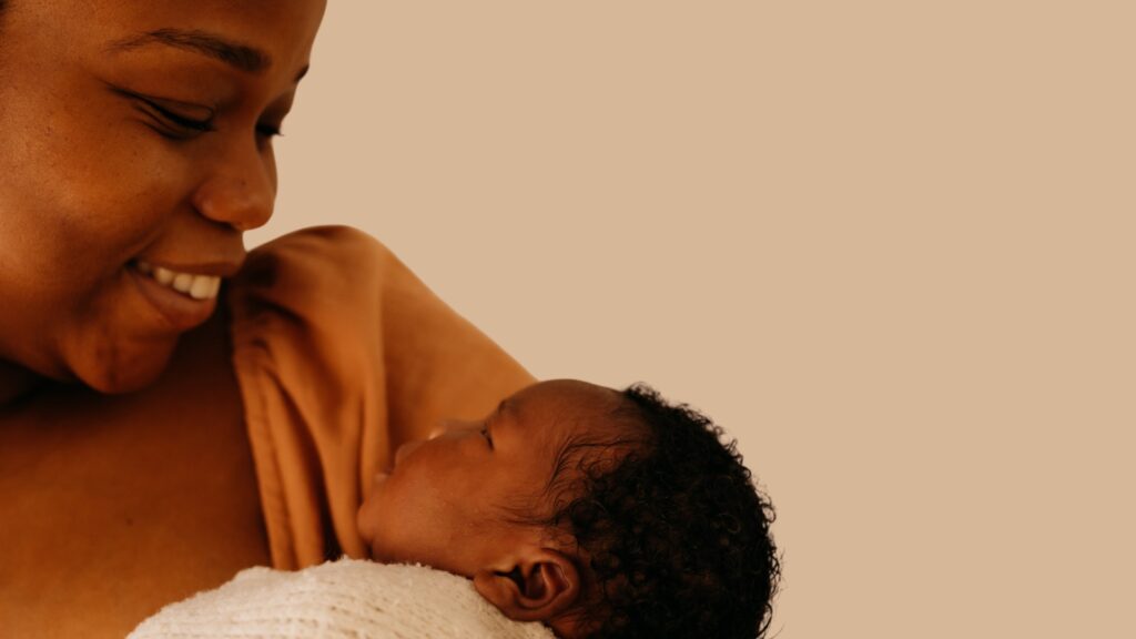 A Black woman smiles as she gazes at an infant she cradles in her arms.