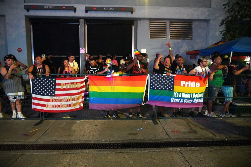 A crowd stands behind a barrier on the sidewalk displaying U.S. and Pride flags with red, orange, yellow, green, blue, and violet stripes and stars. Two of the flags say: “Pride. It’s a right I defend over and over again.”