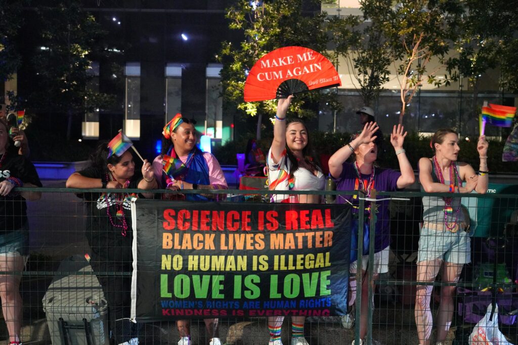 A small group of people stands behind a barrier waving rainbow-colored flags and holding up a red fan that reads “Make Me Cum Again.” On the barrier, a flag reads, “Science Is Real, Black Lives Matter, No Human Is Illegal, Love Is Love, Women’s Rights Are Human Rights, and Kindness Is Everything.”