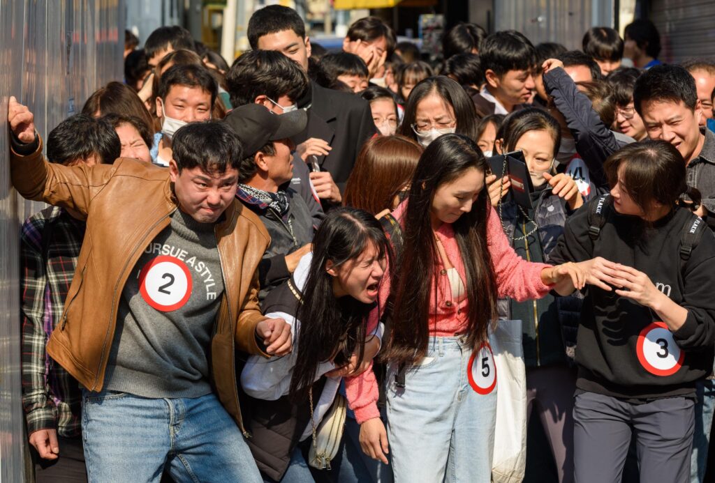 A group of people with dark hair wear casual jeans, T-shirts, and jackets and appear crushed together in a struggle to stand upright. Some wear round white stickers with black numbers on them.