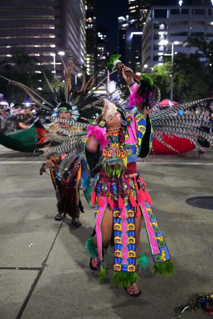 A person clad in blue, green, pink, red, and yellow ceremonial clothing and wearing a feathered headdress dances while blowing into a conch shell on a city street at night.