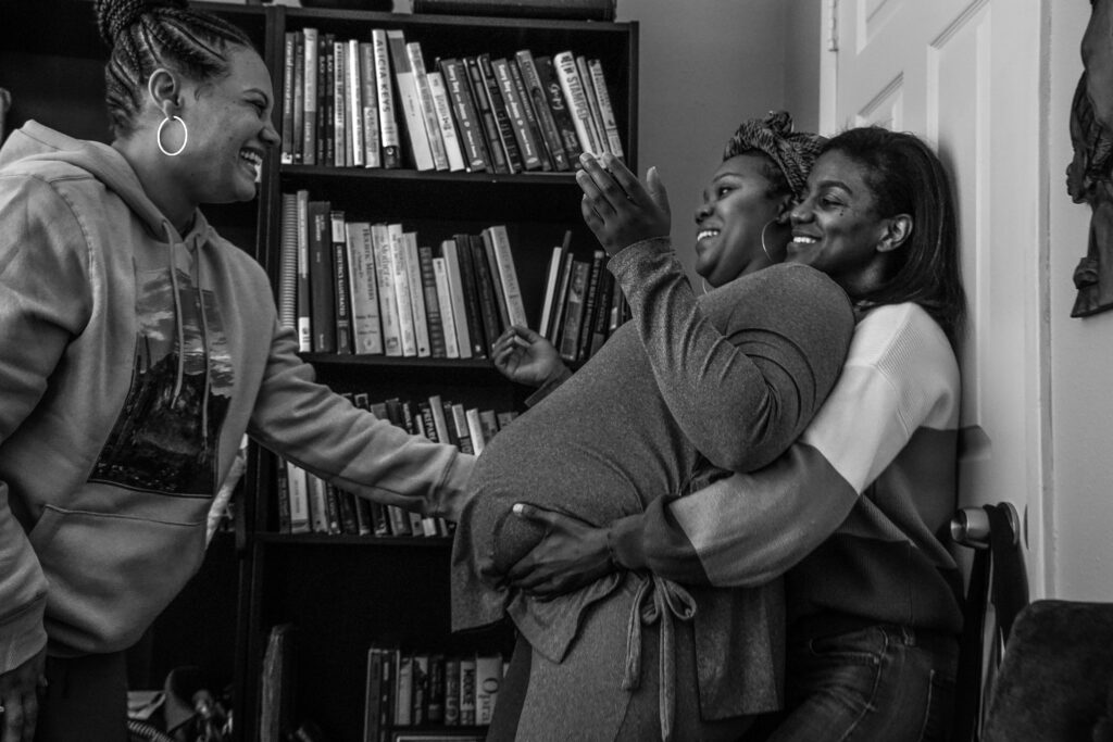 A black-and-white photograph shows three Black women standing in front of a bookshelf. One woman, pregnant, leans her back against another, who reaches around to cradle her baby bump. The third reaches out to touch her belly.