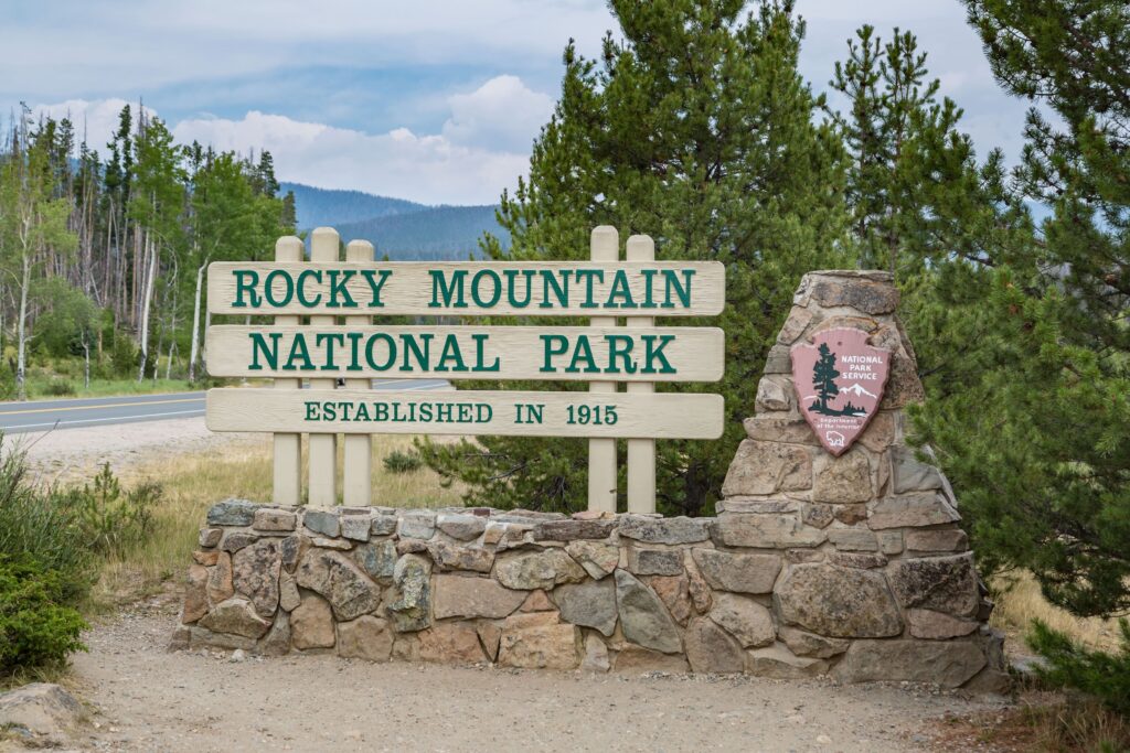 Next to a tree-lined road, a light-colored wooden sign with green letters stands atop a stone wall. It says “Rocky Mountain National Park, Established in 1915.”
