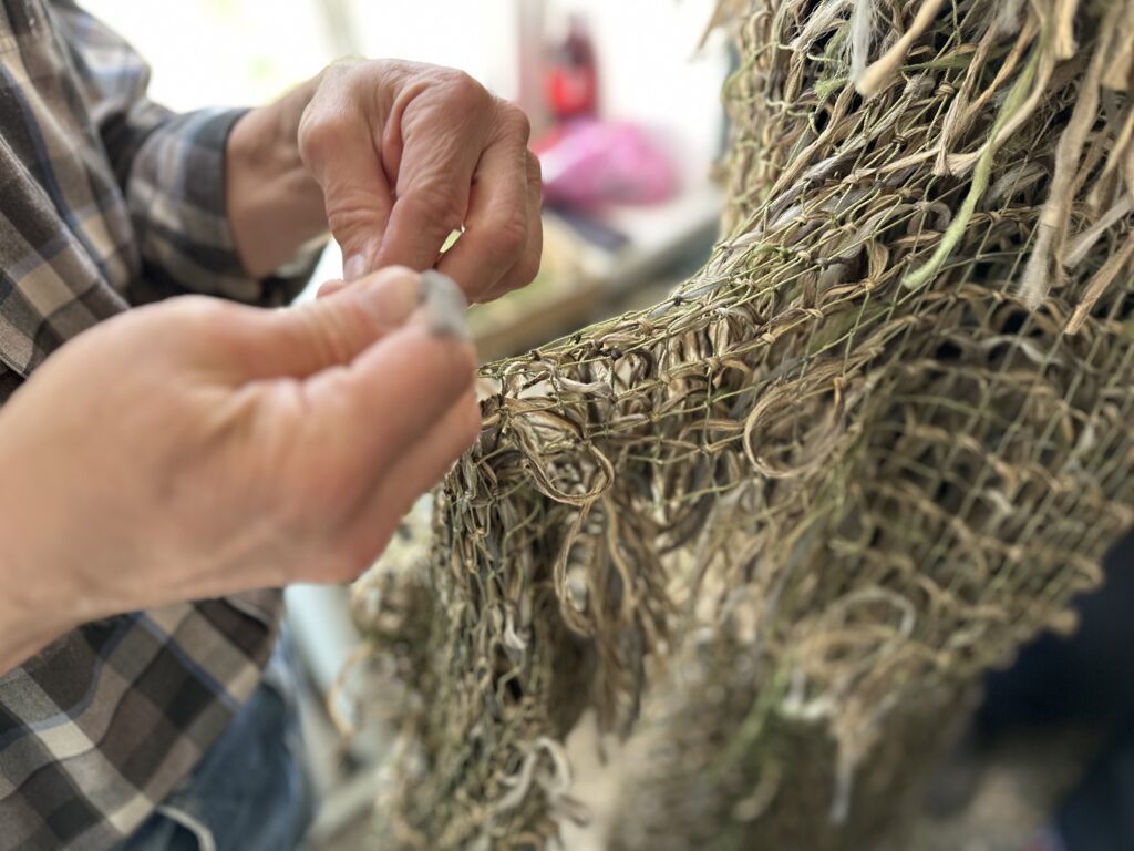A pair of hands belonging to someone wearing a plaid shirt weaves strips of olive-green fabric into a net.