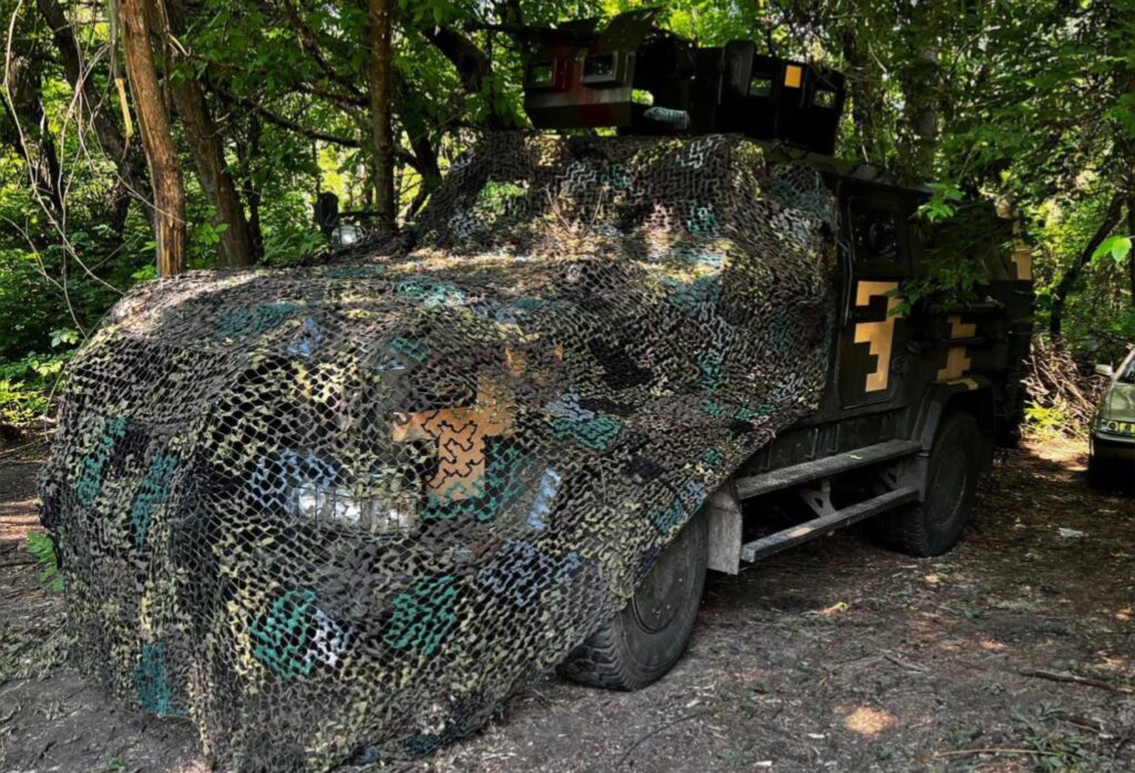 A dark-green colored truck with yellow markings on its side and a rocket launcher mounted on top sits beneath a grove of leafy, green trees. The front of the truck is covered with loose netting mottled with various shades of green.