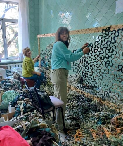 In a brightly lit room with mint-green tiled walls, two women work at a large loom to weave loose netting of mottled black, yellow, green, and gray colors. On the floor at their feet are piles of fabric strips from which the netting is woven.