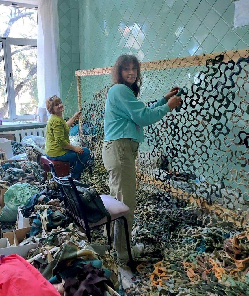 In a brightly lit room with mint-green tiled walls, two women work at a large loom to weave loose netting of mottled black, yellow, green, and gray colors. On the floor at their feet are piles of fabric strips from which the netting is woven.