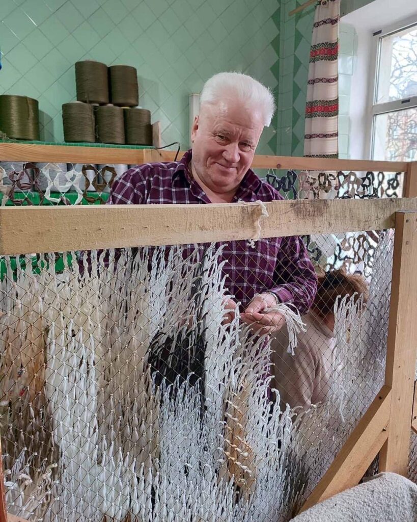 In a room with a window and mint-green tiled walls, a white-haired man wearing a plaid shirt stands at a large wooden loom weaving netting made from strips of white cloth. He smiles slightly.