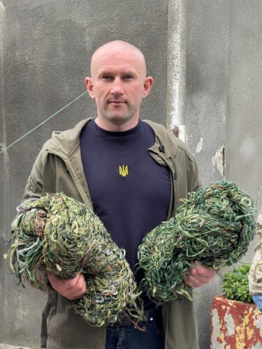 A man with a shaved head, an olive green sweatshirt, and a dark T-shirt with a small, yellow insignia on it holds two bundles of greenish netting.
