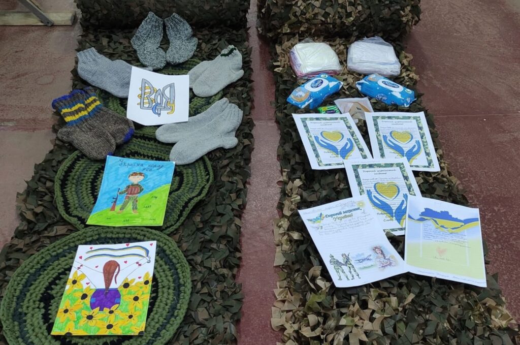 Children's drawings, hand-knit mittens, and other small items lie on top of two narrow strips of dark green netting.