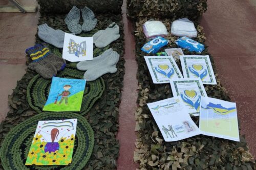 Children's drawings, hand-knit mittens, and other small items lie on top of two narrow strips of dark green netting.