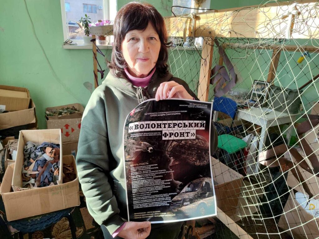 A dark-haired woman wearing an olive green sweatshirt holds up a poster with Cyrillic letters on it. Behind her are piles of cardboard boxes full of strips of fabric.
