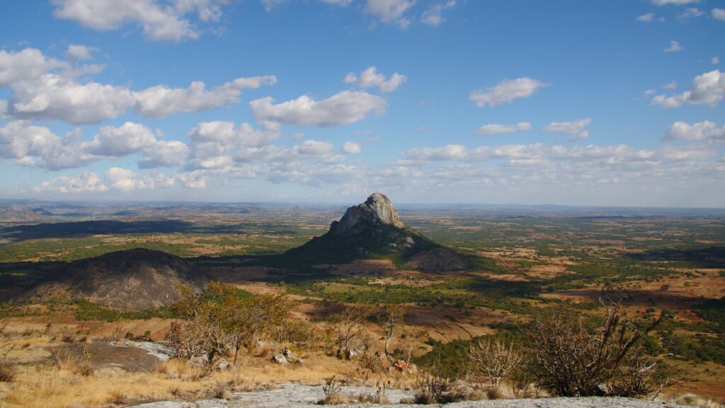 A tall rock structure rises in the middle of a beige and green plain beneath a blue sky dfotted with wispy white clouds.
