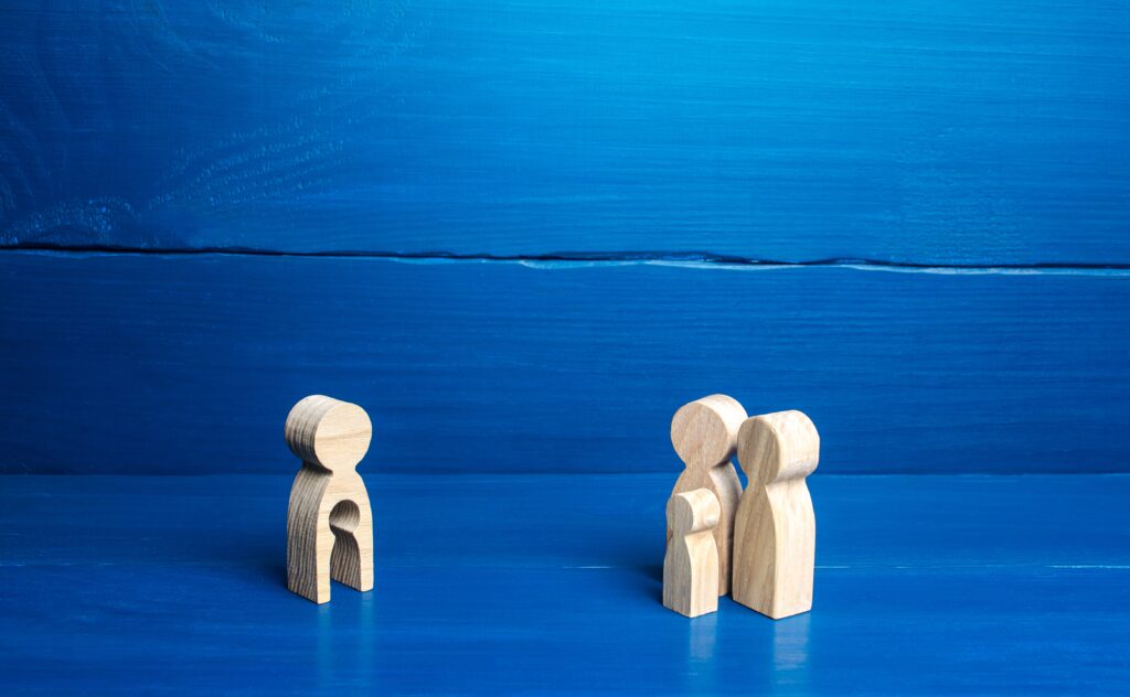 Four person-shaped wooden figurines stand on wood surface that has been painted bright blue. On the right, three cluster together representing parents and a child. On the left, one with a child-shaped cut out stands alone.