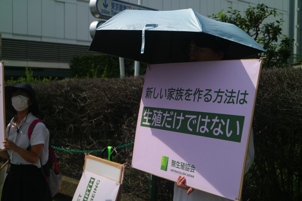 A person holds an umbrella in one hand and a green and white sign covering their face. The sign has Japanese writing and at the bottom says in English: “Antinatalism Japan.” To the person’s right stands a second person wearing a hat and surgical mask.