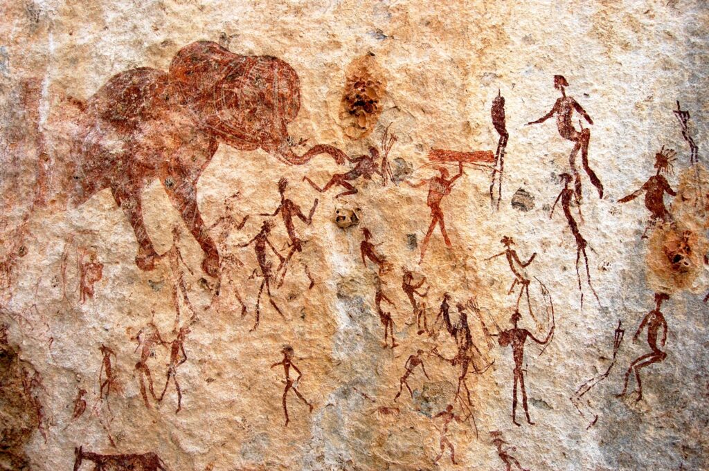 Rust-colored paintings on a beige rock surface depict ancient humans hunting large animals.