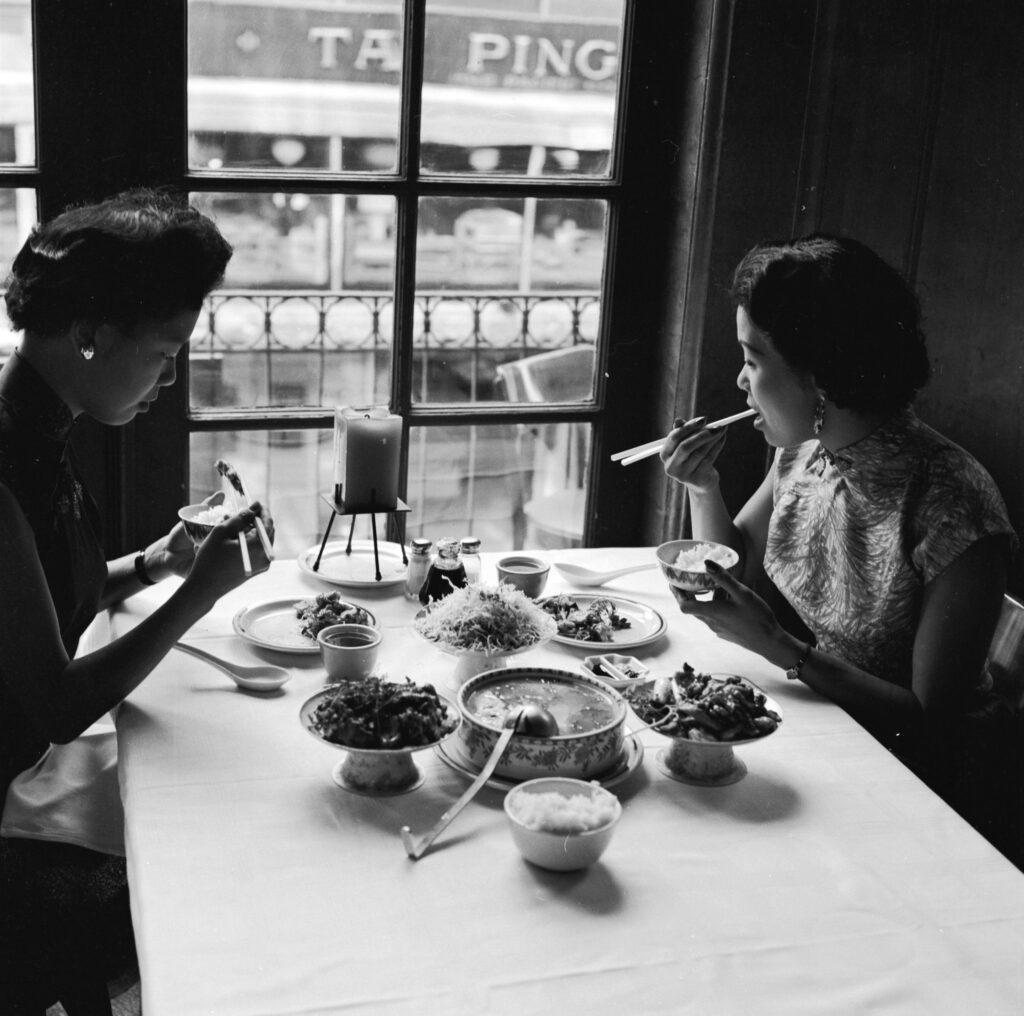 A black-and-white photograph depicts two women sitting by a window in a restaurant. Plates and bowls of food sit on the table as both women use chopsticks to eat from small bowls they hold.
