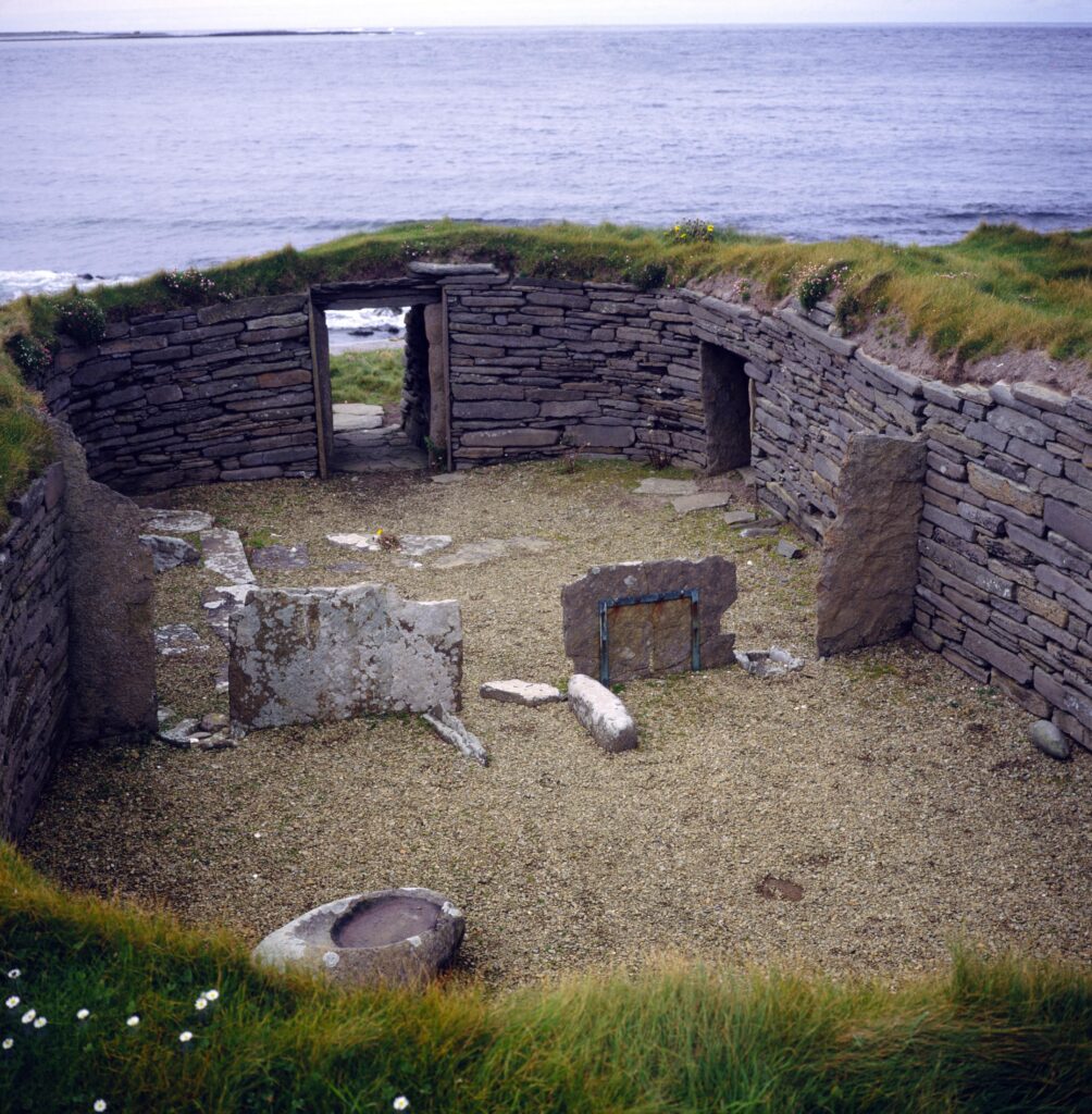 The remains of an ancient stone structure are located amid green grasses beside the ocean.