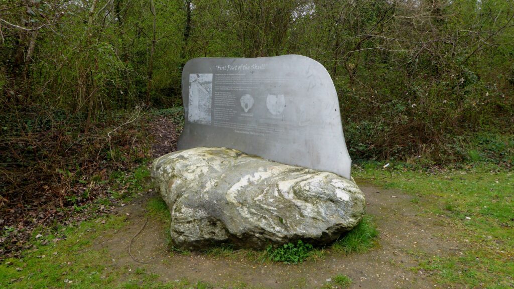A silver-colored metallic monument with writing inscribed on it is affixed to a large stone that sits on a patch of green grass beside thin trees and foliage.
