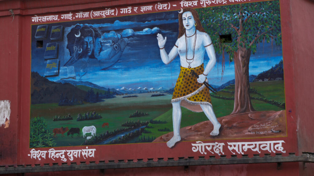 A colorful mural depicts a large, shirtless human figure beside a tree. Cows graze in a field in the background.