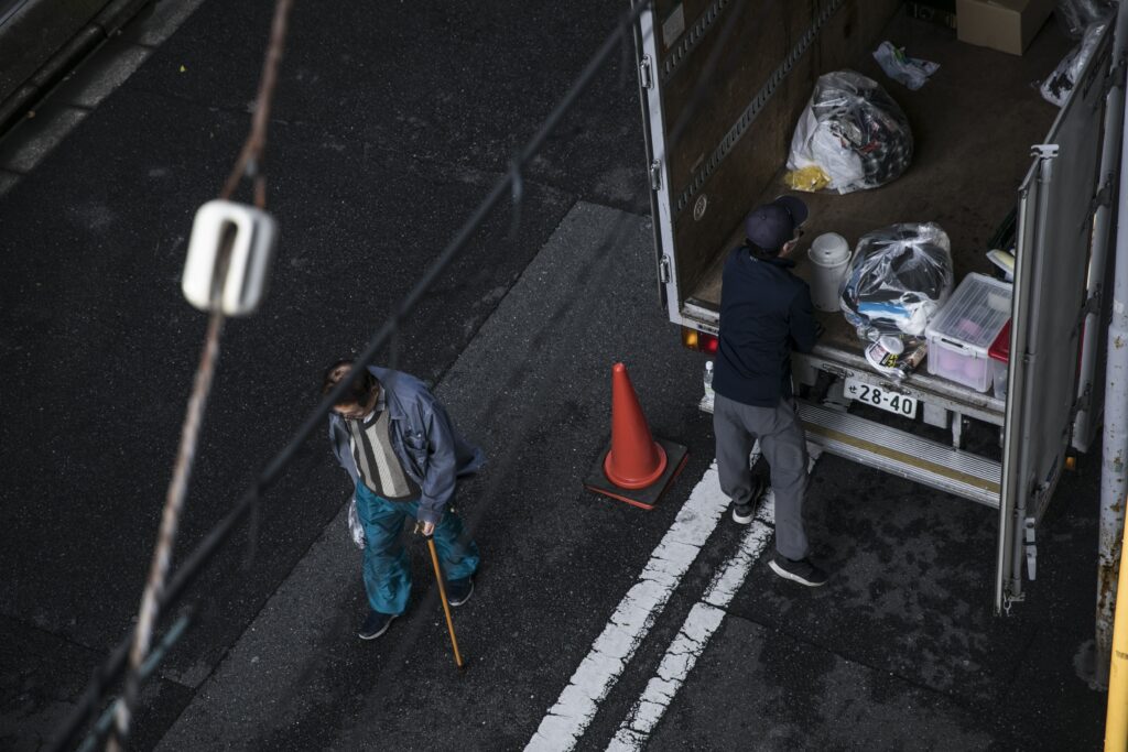 From an overhead angle, a photo shows an older person walking down a street with a cane while a second person loads bags of personal items into a truck.