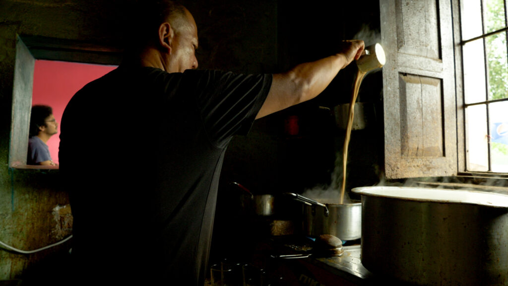 A person in a black shirt standing before an open window in a kitchen raises their arm into the air to pour a stream of light-brown liquid in a boiling pot.