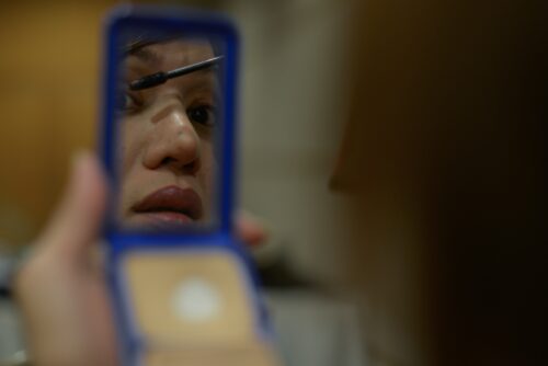 Part of a face is reflected in a handheld compact mirror as a person applies mascara.