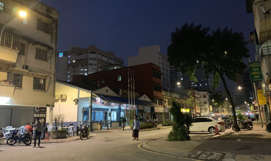 On an urban street corner with parked cars and apartment buildings, people sit outside and walk in the evening.