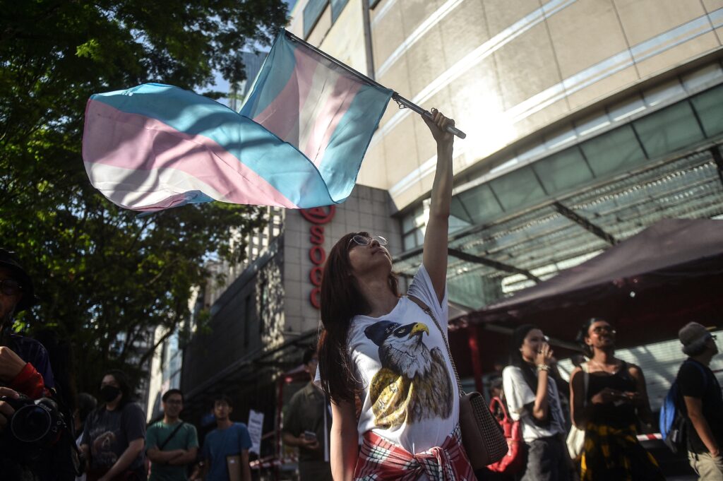 A person with long hair and a T-shirt with an eagle on it holds aloft a pink, white, and blue trans pride flag, which flutters in the breeze as sunlight glints on a nearby building. Other people look on.