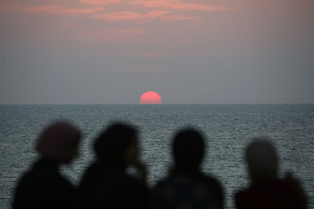 Four silhouetted people stand on a shoreline watching a vivid pink sun set over a calm, expansive ocean under a hazy, pastel sky.